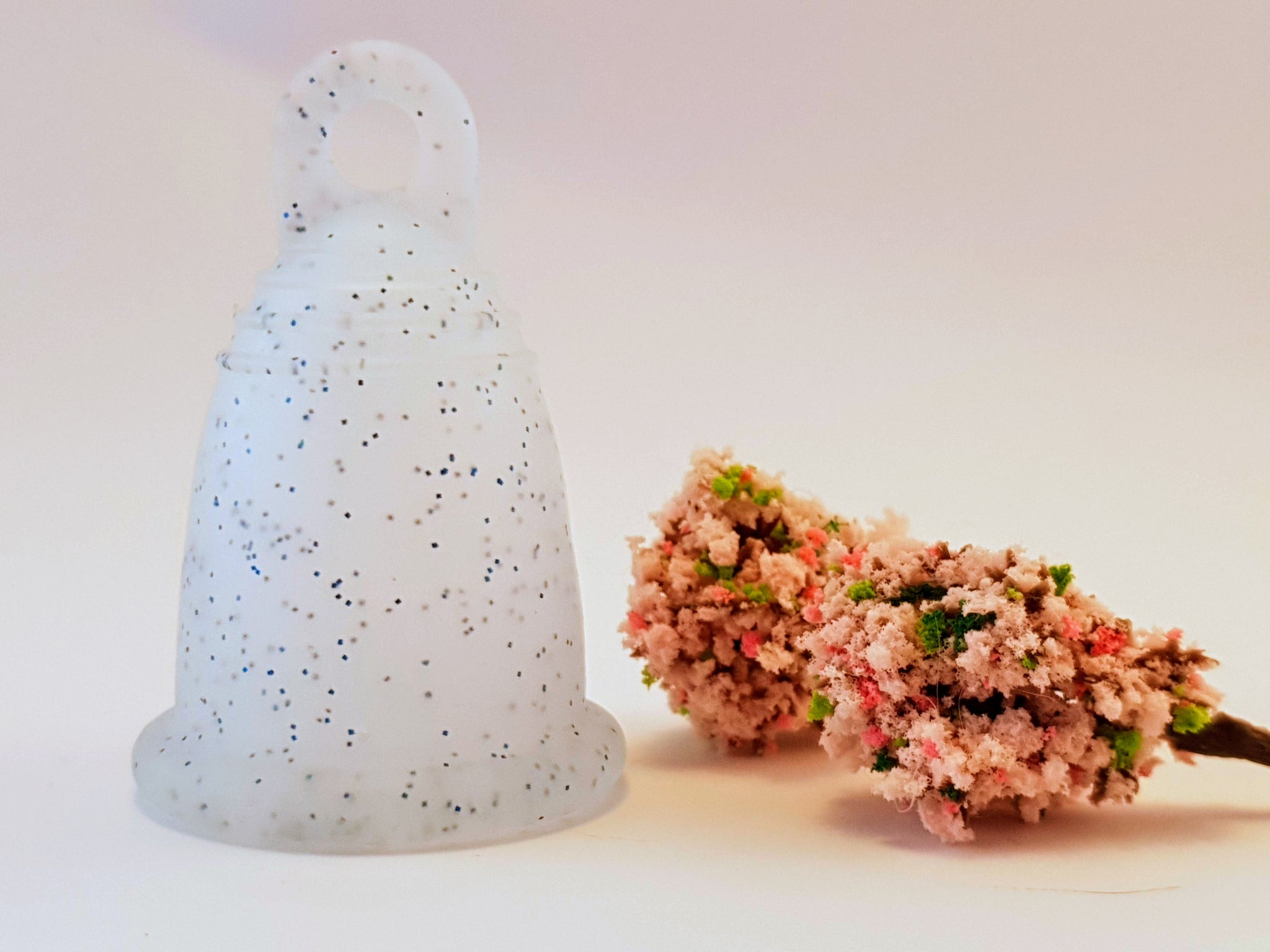 blue glitter menstrual cup, white background and pink flowers, classic Meluna period cup will change the way you feel about your period forever. This menstrual cup is made from reusable soft silicone, the perfect eco-choice for sister's who want a safe, healthy, non toxic, latex free, BPA free גביעונית מחזור או כוס לווסת הכי טובה ונוחה בארץ
